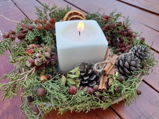 A square white candle is surrounded by a festive wreath made of pinecones, cinnamon sticks, acorns, dried berries, and slices of dried orange. The arrangement sits on a wooden surface.