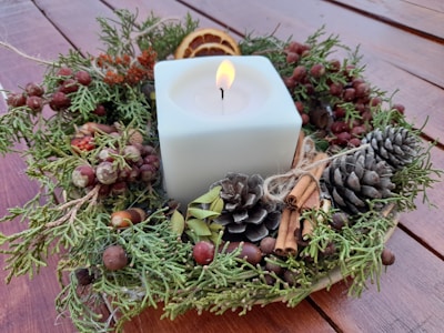 A square white candle is surrounded by a festive wreath made of pinecones, cinnamon sticks, acorns, dried berries, and slices of dried orange. The arrangement sits on a wooden surface.