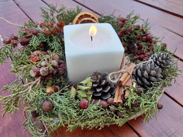 A square white candle is surrounded by a festive wreath made of pinecones, cinnamon sticks, acorns, dried berries, and slices of dried orange. The arrangement sits on a wooden surface.