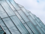 Rows of glass panels form a series of modern architectural structures, with a focus on their sleek, angular design. The panels are transparent and reflect the light, giving a sense of openness and space against a backdrop of a partly cloudy sky.