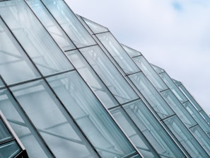 Rows of glass panels form a series of modern architectural structures, with a focus on their sleek, angular design. The panels are transparent and reflect the light, giving a sense of openness and space against a backdrop of a partly cloudy sky.
