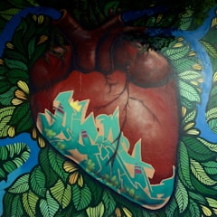 A mural features a realistic depiction of a human heart enveloped by vibrant graffiti art, surrounded by lush foliage and blue accents. The heart's veins and texture are detailed, contrasting with the stylized green and blue graffiti that integrates natural and urban elements.