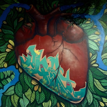 A mural features a realistic depiction of a human heart enveloped by vibrant graffiti art, surrounded by lush foliage and blue accents. The heart's veins and texture are detailed, contrasting with the stylized green and blue graffiti that integrates natural and urban elements.