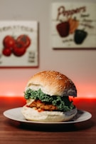 A hamburger with a sesame seed bun is placed on a white plate. The burger includes a crispy patty, fresh green lettuce, and a hint of red sauce. In the background, there are decorative images of vegetables on the wall, and a warm red light creates a cozy ambiance.