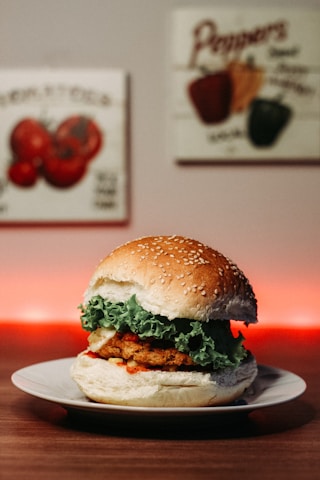 A hamburger with a sesame seed bun is placed on a white plate. The burger includes a crispy patty, fresh green lettuce, and a hint of red sauce. In the background, there are decorative images of vegetables on the wall, and a warm red light creates a cozy ambiance.