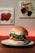 A hamburger with a sesame seed bun is placed on a white plate. The burger includes a crispy patty, fresh green lettuce, and a hint of red sauce. In the background, there are decorative images of vegetables on the wall, and a warm red light creates a cozy ambiance.