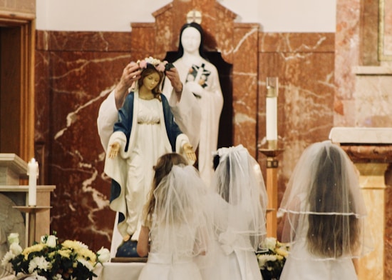A religious ceremony with a statue of the Virgin Mary decorated with a floral crown. The statue is surrounded by young girls wearing white dresses and veils, possibly participating in a First Communion or May Crowning event. The setting includes candles and flower arrangements, with a marble backdrop featuring another religious statue.
