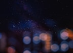 A night sky filled with stars and the Milky Way galaxy, combined with bokeh effect lights in the foreground, creating a blend of cosmic and urban elements.