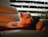 A small white dog is lounging on a cozy, cream-colored leather couch. Ambient lighting casts a warm, orange glow over the scene, creating a relaxed and peaceful atmosphere. A dark cushion supports the dog, and subtle fairy lights are visible behind a striped curtain, adding a touch of whimsy. Potted plants can be seen along the edge of the frame, contributing to a homey setting.