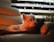 A small white dog is lounging on a cozy, cream-colored leather couch. Ambient lighting casts a warm, orange glow over the scene, creating a relaxed and peaceful atmosphere. A dark cushion supports the dog, and subtle fairy lights are visible behind a striped curtain, adding a touch of whimsy. Potted plants can be seen along the edge of the frame, contributing to a homey setting.