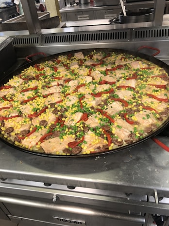 A large pan filled with a vibrant and colorful dish, featuring chunks of fish, bright yellow corn, green peas, red peppers, and sliced sausage. The pan is situated on a metal stovetop in a kitchen setting.