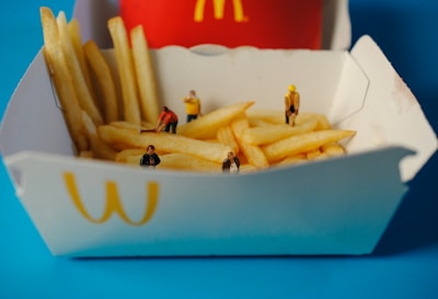 A box of French fries with miniature figurines of people placed among the fries. The scene includes various figures engaged as if they are working or interacting with the fries. In the background, a red container with a yellow letter logo is visible, set against a bright blue background.