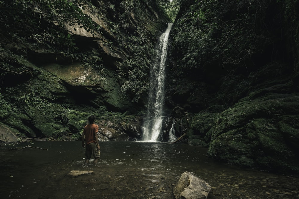 man in red jacket standing on brown rock near waterfalls during daytime