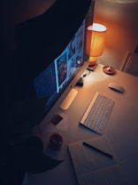 A cozy workspace featuring a desktop computer displaying a gallery of images, illuminated by a warm table lamp. The setup includes a wireless keyboard, mouse, open notebook with handwritten notes, and a small candle on the desk. A plant partially obscures the view on one side, adding a touch of greenery to the scene.