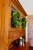 A wooden cabinet with glass doors adorned with lush green wreaths. Several small decorative objects, including wooden pepper mills and small potted plants, sit on the cabinet surface. The cabinet is set against a background featuring a light-colored brick wall.