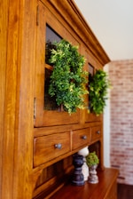 A wooden cabinet with glass doors adorned with lush green wreaths. Several small decorative objects, including wooden pepper mills and small potted plants, sit on the cabinet surface. The cabinet is set against a background featuring a light-colored brick wall.