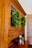 A wooden cabinet with glass doors adorned with lush green wreaths. Several small decorative objects, including wooden pepper mills and small potted plants, sit on the cabinet surface. The cabinet is set against a background featuring a light-colored brick wall.