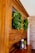 A wooden cabinet with glass doors adorned with lush green wreaths. Several small decorative objects, including wooden pepper mills and small potted plants, sit on the cabinet surface. The cabinet is set against a background featuring a light-colored brick wall.