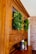 A wooden cabinet with glass doors adorned with lush green wreaths. Several small decorative objects, including wooden pepper mills and small potted plants, sit on the cabinet surface. The cabinet is set against a background featuring a light-colored brick wall.