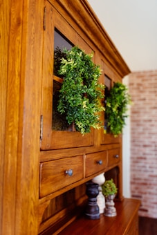 A wooden cabinet with glass doors adorned with lush green wreaths. Several small decorative objects, including wooden pepper mills and small potted plants, sit on the cabinet surface. The cabinet is set against a background featuring a light-colored brick wall.