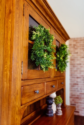 A wooden cabinet with glass doors adorned with lush green wreaths. Several small decorative objects, including wooden pepper mills and small potted plants, sit on the cabinet surface. The cabinet is set against a background featuring a light-colored brick wall.