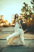 A woman in a flowing wedding dress is captured walking on a road with the setting sun in the background. Her hair and gown move dramatically in the wind, adding a sense of motion to the scene. The ambient light casts a warm glow over the image, highlighting the dress's intricate details and the tattoos on her arm.