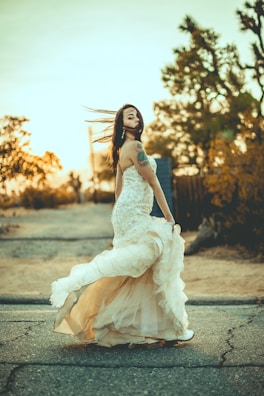 A woman in a flowing wedding dress is captured walking on a road with the setting sun in the background. Her hair and gown move dramatically in the wind, adding a sense of motion to the scene. The ambient light casts a warm glow over the image, highlighting the dress's intricate details and the tattoos on her arm.