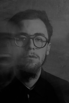 A monochrome portrait of a man with glasses and a beard, captured in a dreamy or ghostly double exposure effect. The image conveys a sense of mystery and introspection with its blurred and sharp contrasts.