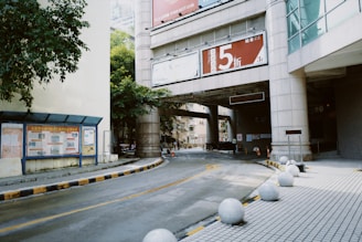 An urban scene with a curved road leading under a bridge connected to a modern building. The road is lined with round concrete spheres, likely for pedestrian safety. A large sign with a red background and bold text is prominently displayed on the bridge. The surrounding architecture includes high-rise structures and greenery peeks through the gaps. Signs and boards are present along the sidewalk.
