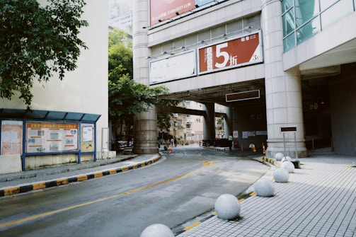 An urban scene with a curved road leading under a bridge connected to a modern building. The road is lined with round concrete spheres, likely for pedestrian safety. A large sign with a red background and bold text is prominently displayed on the bridge. The surrounding architecture includes high-rise structures and greenery peeks through the gaps. Signs and boards are present along the sidewalk.