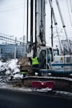 A construction site featuring a large drilling machine with two workers in high-visibility vests. The machinery is positioned beside a snow-covered road, and there are piles of excavated earth around. In the background, there are railway tracks and electrical wires, possibly indicating an urban setting.