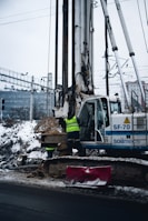 A construction site featuring a large drilling machine with two workers in high-visibility vests. The machinery is positioned beside a snow-covered road, and there are piles of excavated earth around. In the background, there are railway tracks and electrical wires, possibly indicating an urban setting.