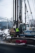 A construction site featuring a large drilling machine with two workers in high-visibility vests. The machinery is positioned beside a snow-covered road, and there are piles of excavated earth around. In the background, there are railway tracks and electrical wires, possibly indicating an urban setting.