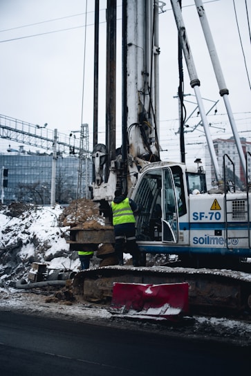 A construction site featuring a large drilling machine with two workers in high-visibility vests. The machinery is positioned beside a snow-covered road, and there are piles of excavated earth around. In the background, there are railway tracks and electrical wires, possibly in
