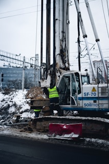 A construction site featuring a large drilling machine with two workers in high-visibility vests. The machinery is positioned beside a snow-covered road, and there are piles of excavated earth around. In the background, there are railway tracks and electrical wires, possibly indicating an urban setting.