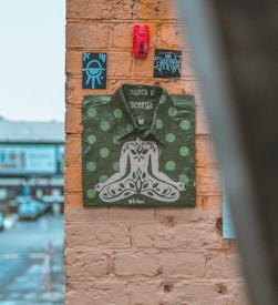 A piece of street art on a brick wall features a painted green and white polka dot shirt with an intricate design forming a seated figure. Above the shirt, there are small black and blue rectangular signs and a red object. The background shows part of an urban street with cars and buildings.