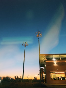 Two small wind turbines are mounted on tall poles near a modern building with large windows. The scene is set during twilight with a clear sky transitioning from deep blue to warm orange near the horizon.