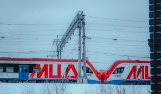A modern train with bold red and blue designs is on a snowy railway track. Overhead power lines and a support structure can be seen above the train. The area is covered in snow, indicating a cold season.