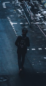 A person walks alone on a dark street, carrying a backpack. The street has wet patches and some markings. Metal railings and caution tape are visible nearby, suggesting construction or restricted access.