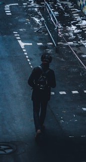 A person walks alone on a dark street, carrying a backpack. The street has wet patches and some markings. Metal railings and caution tape are visible nearby, suggesting construction or restricted access.
