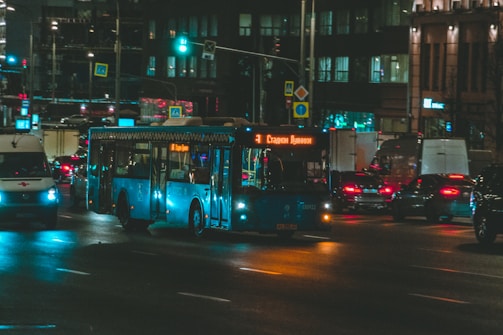 A city street at night with a blue bus in the center, streetlights illuminating the scene. The bus displays a destination in Cyrillic, and there are other vehicles like an ambulance and cars on the road. Buildings with lit windows and neon signs form the background.