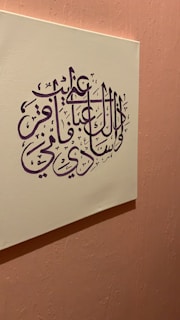 An artwork featuring Arabic calligraphy in deep purple ink set on a cream-colored canvas. The canvas is mounted on a textured, soft pink wall, creating a harmonious contrast between the colors. The calligraphy is intricate and stylized, with elegant curves and lines.