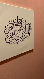 An artwork featuring Arabic calligraphy in deep purple ink set on a cream-colored canvas. The canvas is mounted on a textured, soft pink wall, creating a harmonious contrast between the colors. The calligraphy is intricate and stylized, with elegant curves and lines.