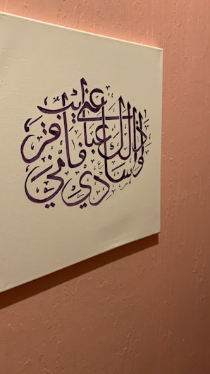 An artwork featuring Arabic calligraphy in deep purple ink set on a cream-colored canvas. The canvas is mounted on a textured, soft pink wall, creating a harmonious contrast between the colors. The calligraphy is intricate and stylized, with elegant curves and lines.