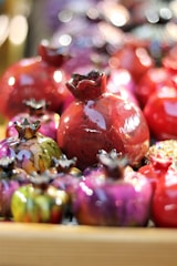 A collection of colorful, glossy ceramic pomegranate sculptures. The sculptures vary in size and color, with vibrant shades of red, purple, pink, and green. The surfaces of the sculptures are reflective, giving them a shiny appearance. They are placed closely together, creating a visually appealing composition.