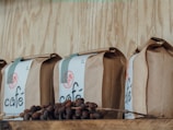 A row of brown paper bags labeled 'café' is displayed on a wooden shelf. In front of the bags, there are coffee cherries still on a branch, adding a rustic and organic feel.