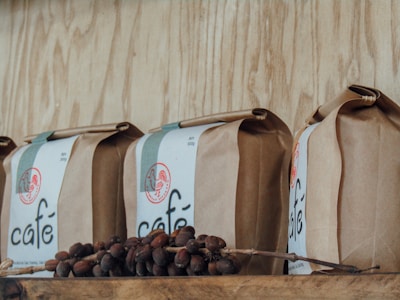 A row of brown paper bags labeled 'café' is displayed on a wooden shelf. In front of the bags, there are coffee cherries still on a branch, adding a rustic and organic feel.