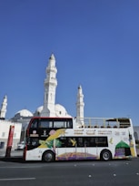 A colorful double-decker tour bus is parked in front of a large white mosque with multiple domes and tall minarets. The bus features vibrant artwork and signage, promoting sightseeing tours in Al Madinah. The background is a clear blue sky, adding to the striking contrast between the white architecture and the bus.