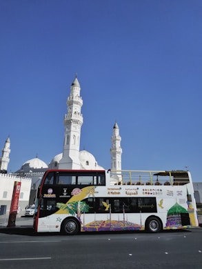 A colorful double-decker tour bus is parked in front of a large white mosque with multiple domes and tall minarets. The bus features vibrant artwork and signage, promoting sightseeing tours in Al Madinah. The background is a clear blue sky, adding to the striking contrast between the white architecture and the bus.