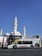 A colorful double-decker tour bus is parked in front of a large white mosque with multiple domes and tall minarets. The bus features vibrant artwork and signage, promoting sightseeing tours in Al Madinah. The background is a clear blue sky, adding to the striking contrast between the white architecture and the bus.